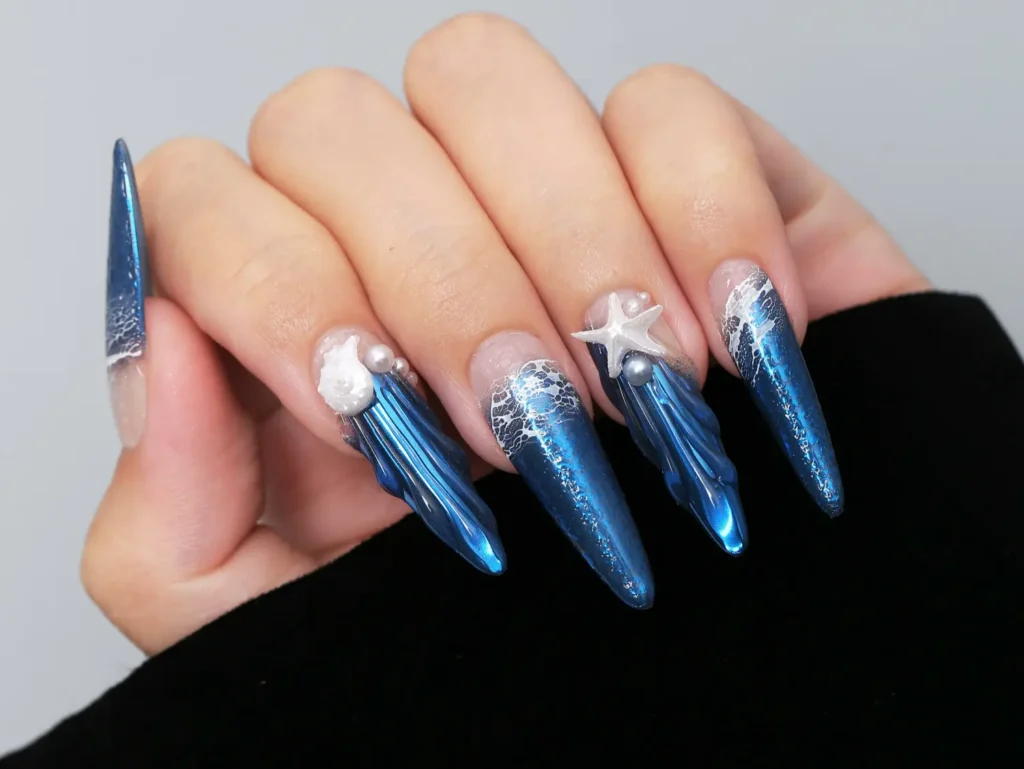 High-end professional salon stiletto nails featuring ocean blue mirror powder with 3D water ripple texture, pearls, and starfish accents