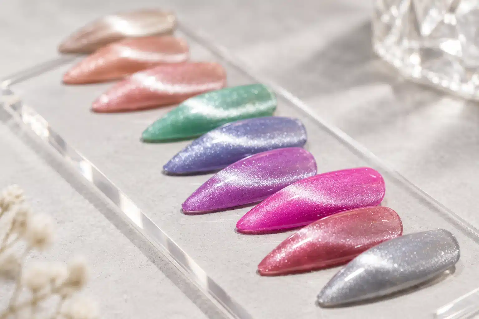 Glittering cat eye nail swatches in pink, green, purple and silver arranged on a clear display stand.