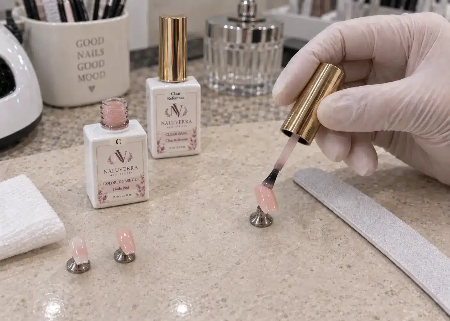 A rounded brush applying nude pink colored base gel to a nail tip beside the matching sample bottle