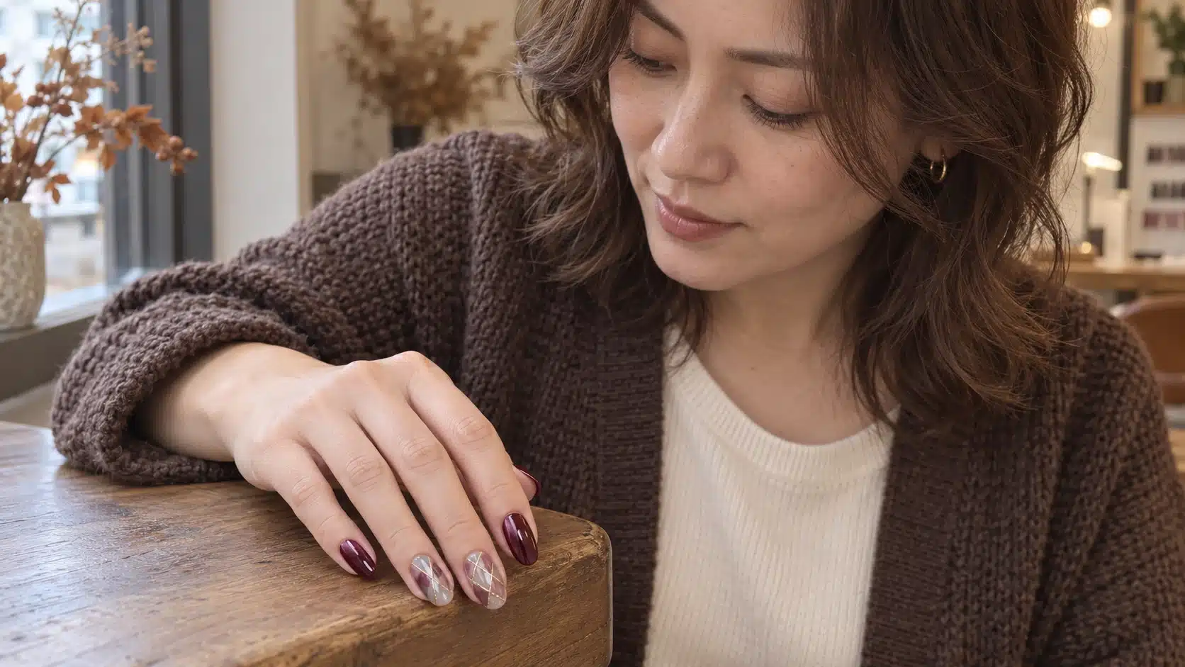 Woman wearing burgundy cat-eye and fine gold-lined plaid nails with a brown cardigan in a nail studio