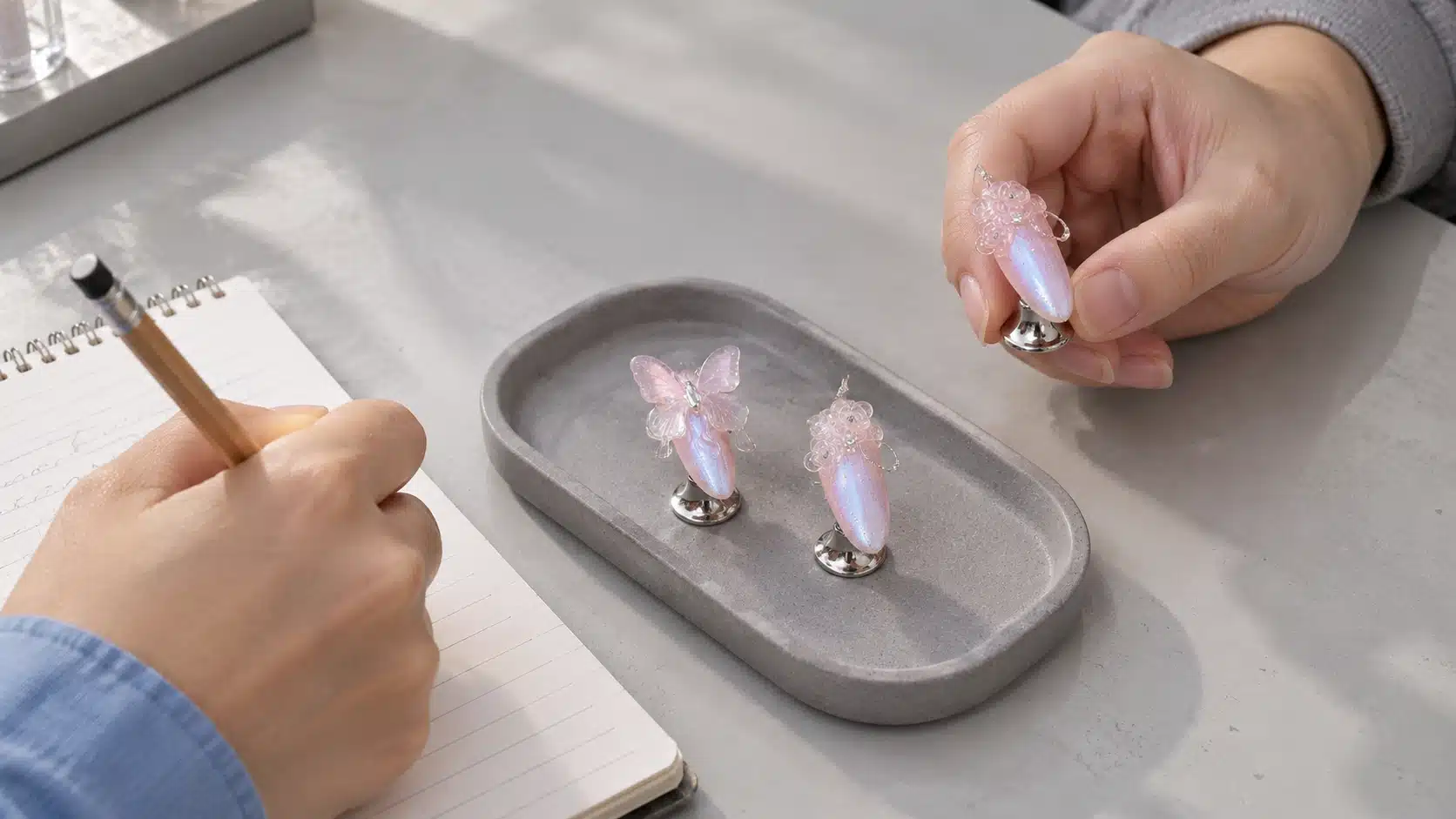 Tabletop Review of Clear Sculpted Nail Samples Hands reviewing clear rose and butterfly nail samples on silver stands beside a notebook