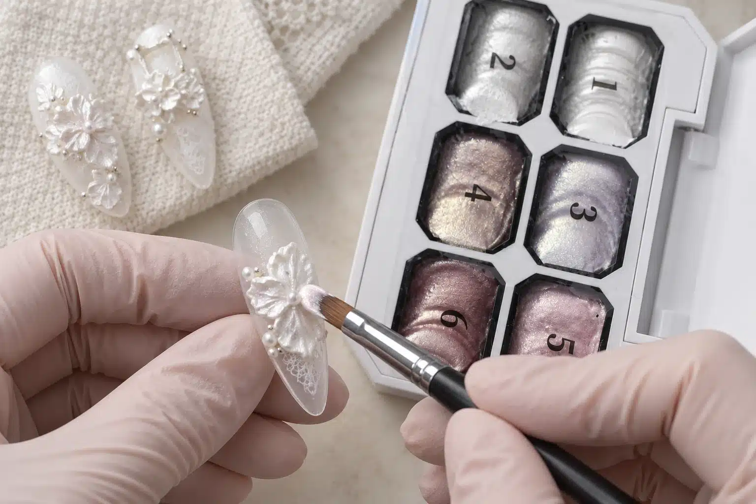Gloved nail technician using a brush to shape a white flower petal on a practice nail tip beside the Dusty Pink Sculpture Gel palette