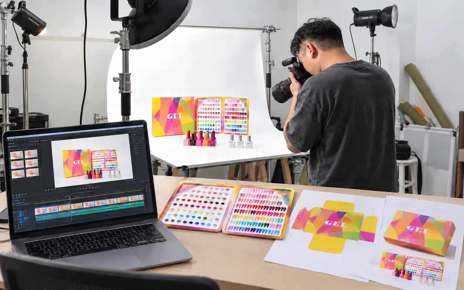 Photographer capturing a gel nail polish collection beside a laptop with a video timeline, packaging proofs and shade charts