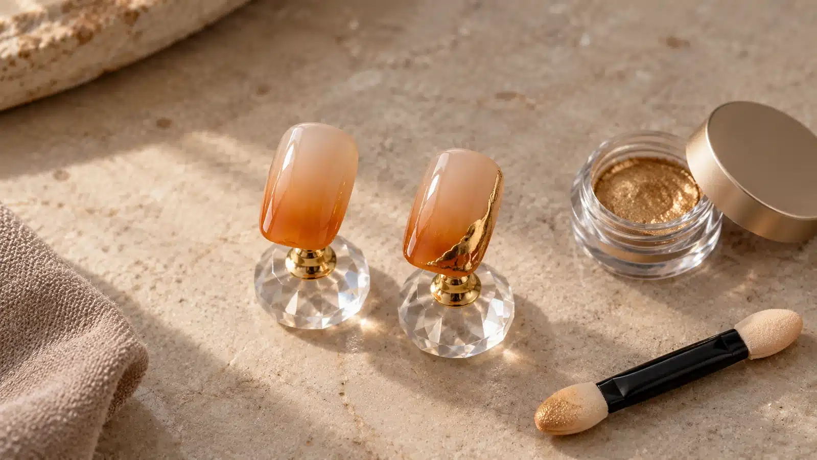 Two caramel ombre nail tips sit on clear stands, one with a gold edge accent, beside gold powder and a foam applicator.