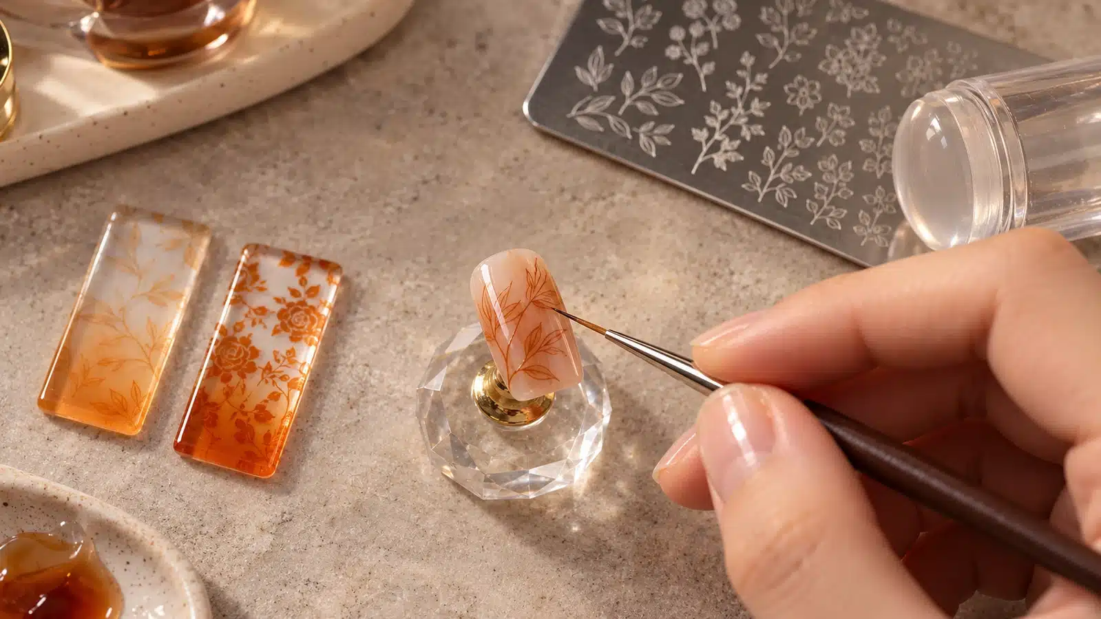 A fine brush touches a leaf-patterned nude nail tip beside amber stamped samples, a metal stamping plate, and a clear stamper.