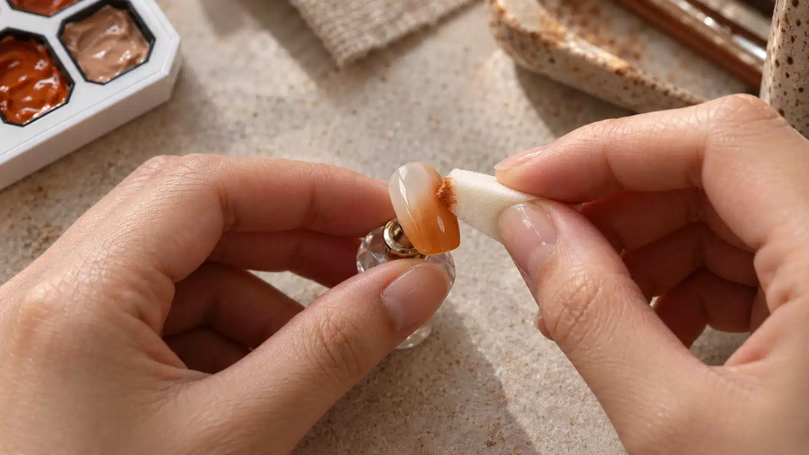 A small sponge touches the right edge of a caramel ombre nail tip held on a magnetic stand