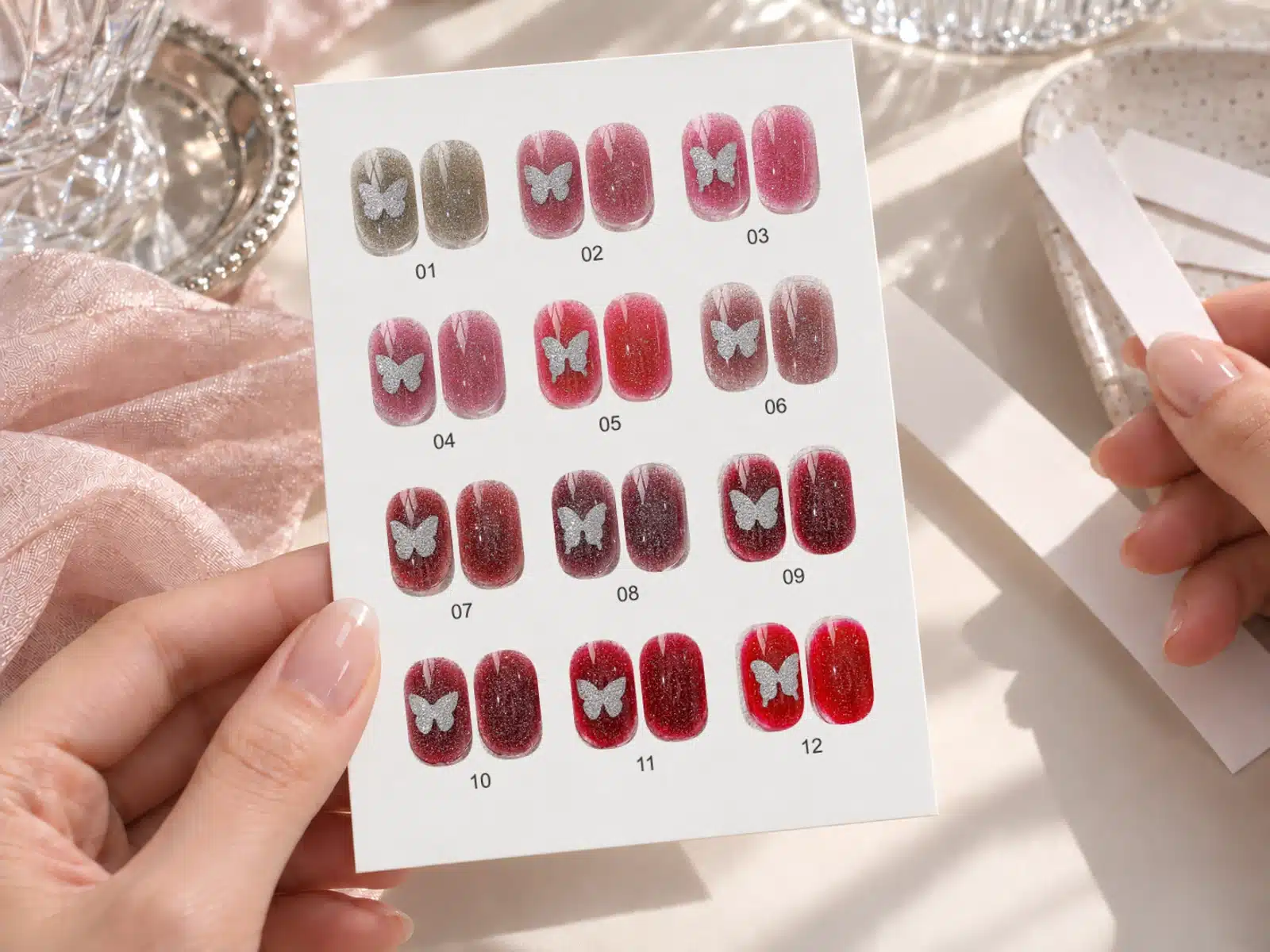 Hands holding twelve numbered pairs of reflective gel nail samples with silver butterfly accents